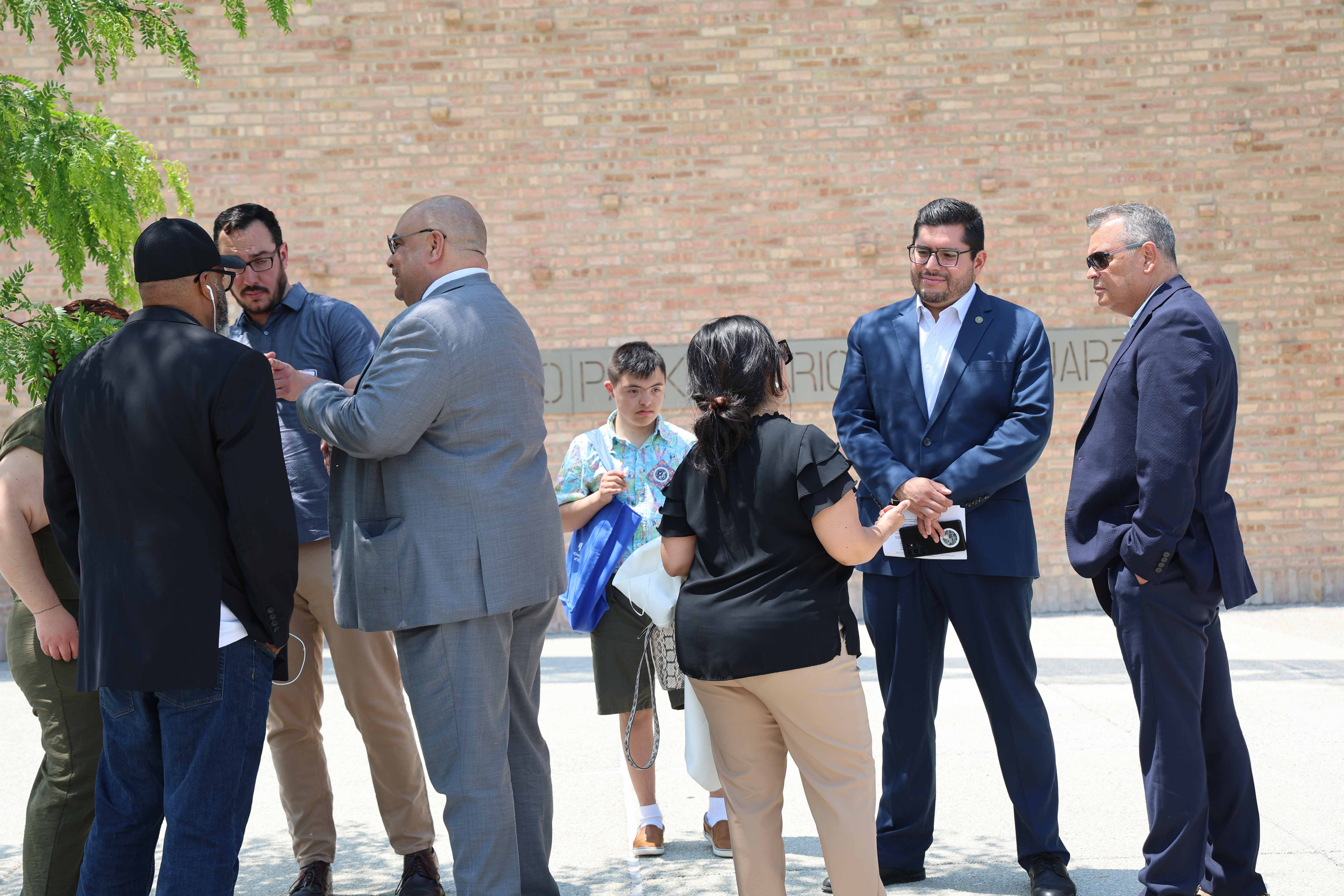 A group of people stand in casual conversation outdoors, in front of a brick wall.